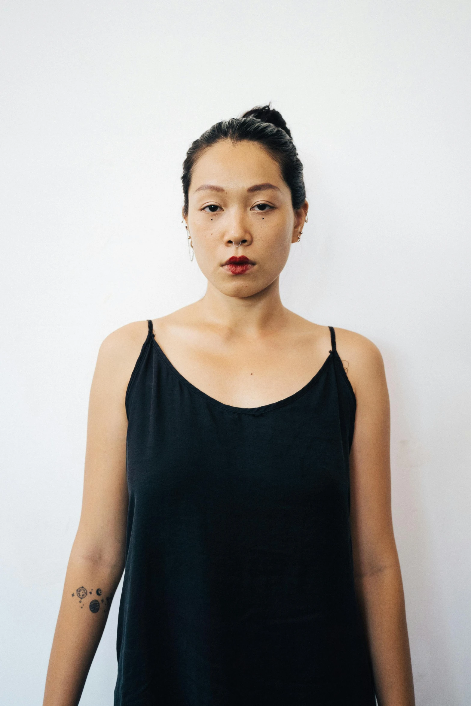 a woman standing in front of a white wall, an album cover, inspired by Ruth Jên, unsplash, she is wearing a black tank top, serious and stern expression, asian descent, black dress : : symmetrical face