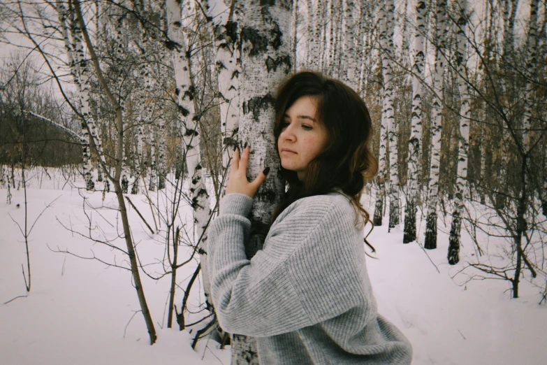 a woman standing next to a tree in the snow, inspired by Elsa Bleda, pexels contest winner, aestheticism, girl with brown hair, wearing a sweater, avatar image, white and grey