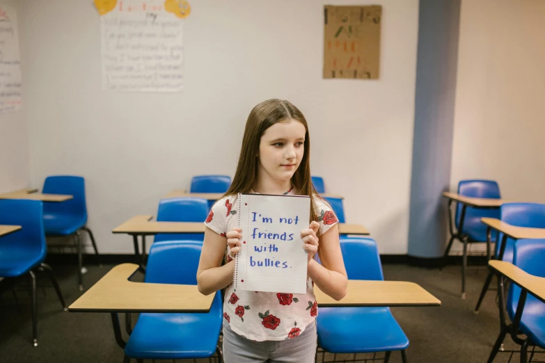 a girl holding a sign in a classroom, trending on unsplash, fan favorite, bullying, high quality photo, blank stare”