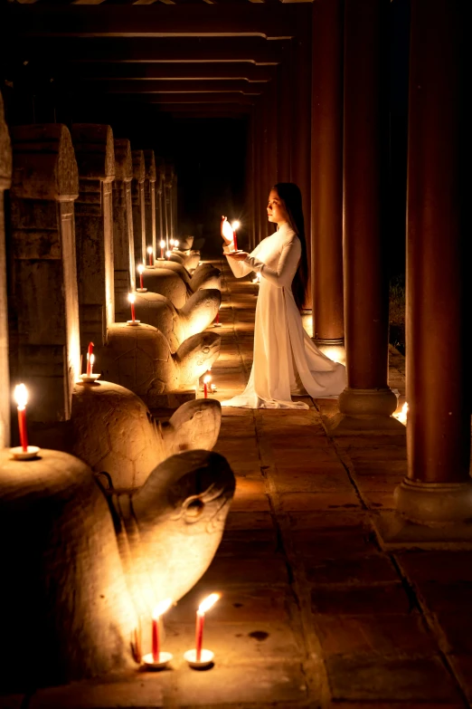 a woman in a long white dress holding a lit candle, a statue, inspired by Steve McCurry, romanesque, angkor, glowing lamps, pillars, warm lighting inside