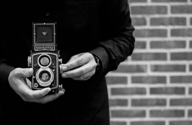 a man holding a camera in front of a brick wall, a black and white photo, art photography, rolleiflex tlr, unsplash photography, old timey, medium format