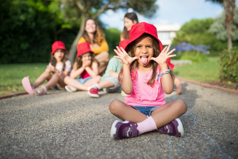 a little girl sitting on the ground with her hands in the air, pexels contest winner, red hat, college girls, long tongue, summer camp