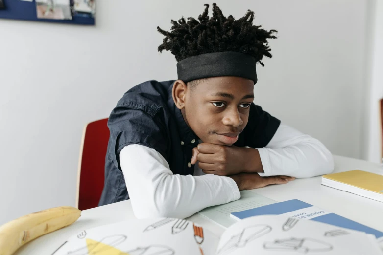 a young boy sitting at a table with a banana, by Barthélemy Menn, pexels contest winner, black teenage boy, sitting in the classroom, thinking pose, on a white table