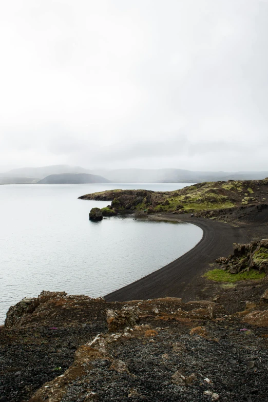 a black sand beach next to a body of water, by Þórarinn B. Þorláksson, lake in the distance, wide views, crater, overcast day