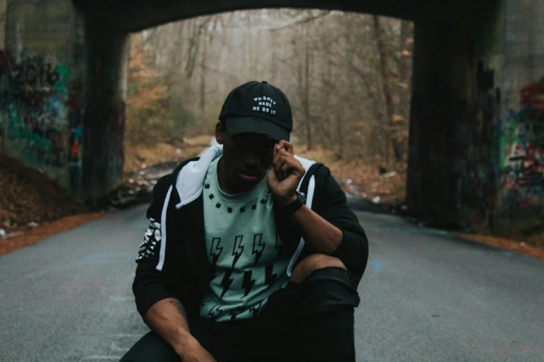 a man sitting on the ground talking on a cell phone, pexels contest winner, graffiti, with rap cap on head, outlive streetwear collection, black and green, near forest