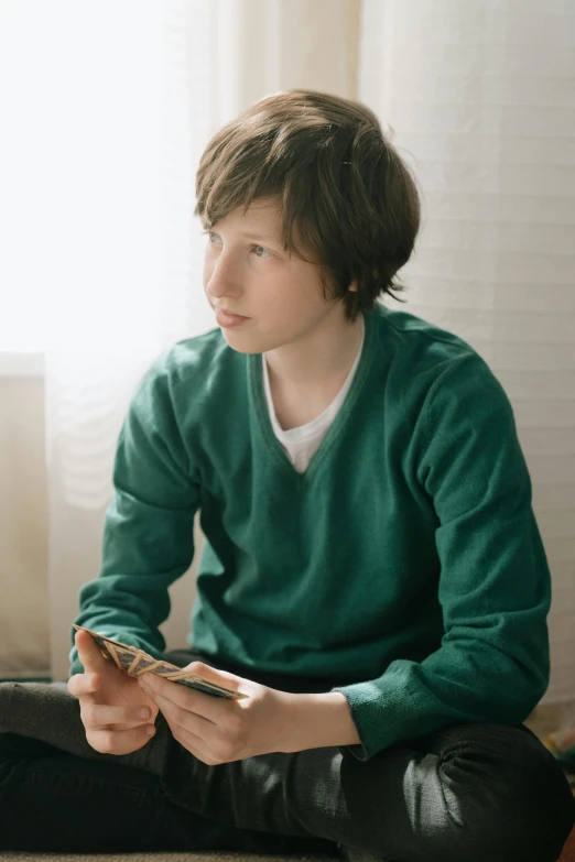 a young man sitting on the floor looking at his cell phone, an album cover, unsplash, hyperrealism, wearing a green sweater, portrait of 1 5 - year - old boy, subreddit / r / whale, hikkikomori