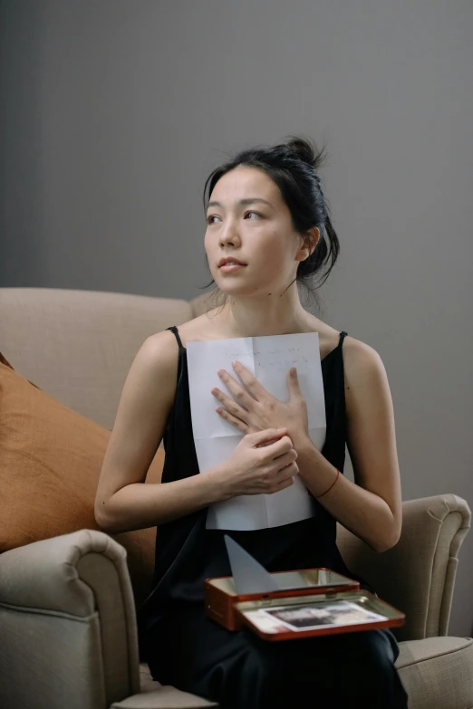 a woman sitting in a chair holding a piece of paper, inspired by Fei Danxu, pexels contest winner, hyperrealism, sitting with wrists together, maggie cheung, indoor scene, holding books