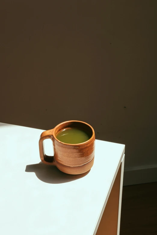 a cup of green tea sitting on top of a white table, by Jessie Algie, terracotta, green ambient light, ignant, moringa juice