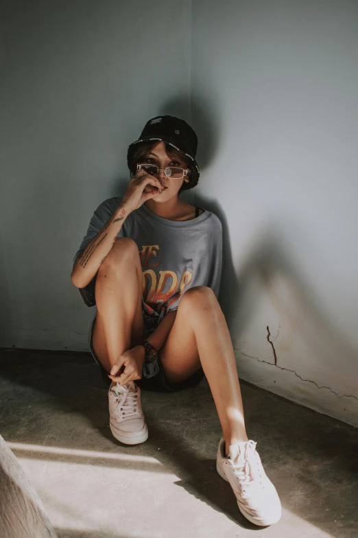 a woman sitting on the ground talking on a cell phone, inspired by Elsa Bleda, trending on pexels, graffiti, tan skin a tee shirt and shorts, standing in corner of room, wearing sunglasses and a cap, portrait androgynous girl