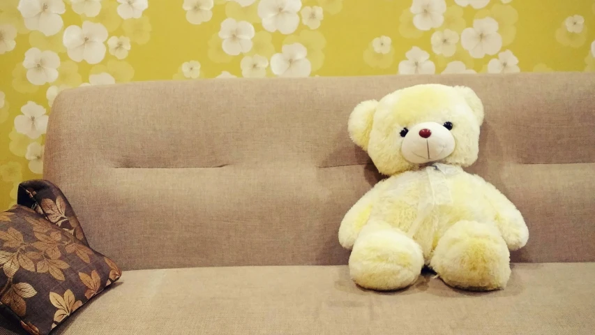 a teddy bear sitting on a couch next to a pillow, yellow wallpaper, at the sitting couch, ross tan, furnished room