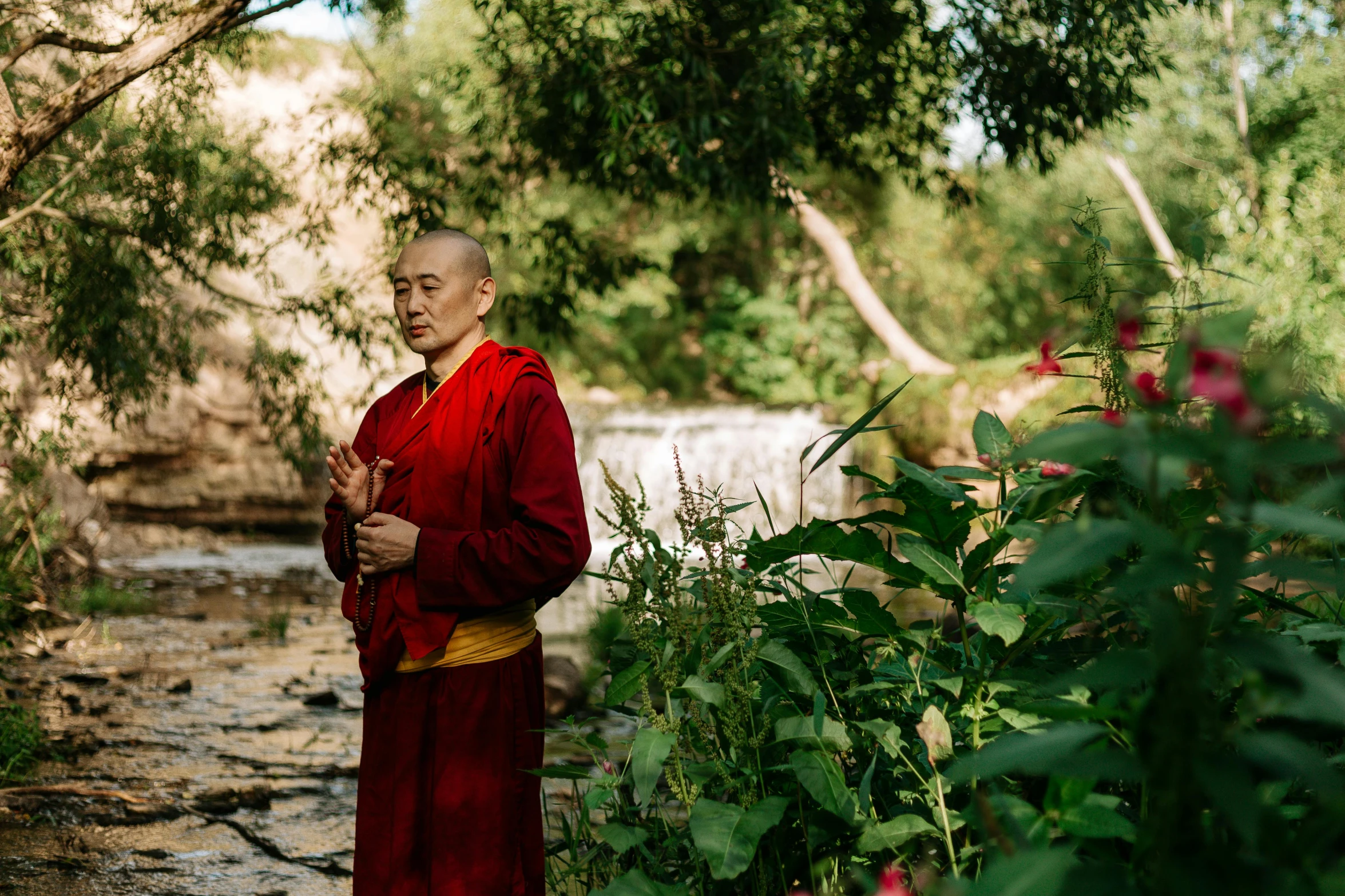 a man in a red robe standing next to a river, a portrait, inspired by Li Di, unsplash, buddhist monk meditating, amongst foliage, taken with sony alpha 9, gong li