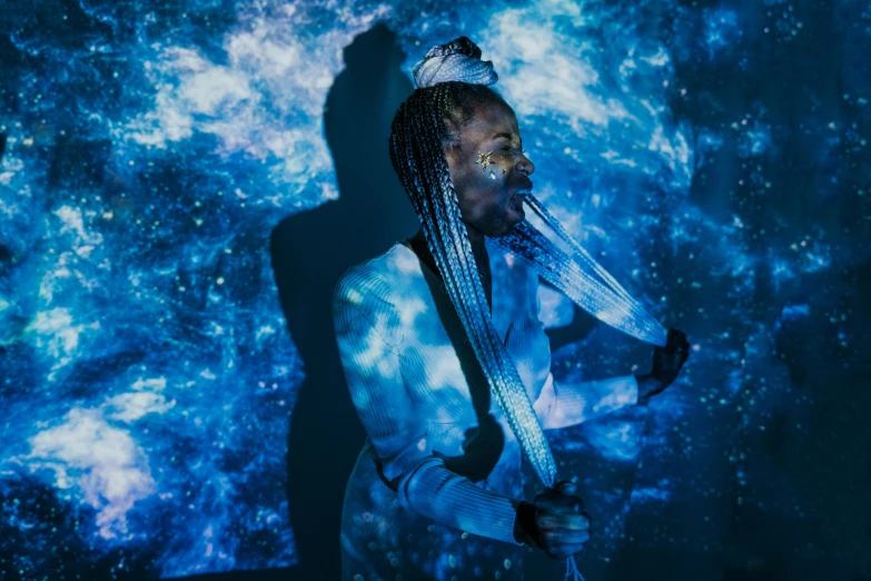 a man with dreadlocks standing in front of a blue background, a hologram, by Julia Pishtar, pexels contest winner, afrofuturism, wearing a dress made of stars, projection design installation, white light shining on her, two long braids blue