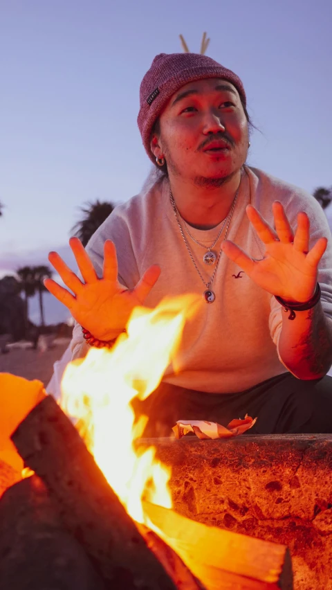 a man sitting next to a fire on a beach, an album cover, by Jessie Alexandra Dick, trending on unsplash, hand gesture, andy milonakis, high quality photo, aboriginal australian hipster