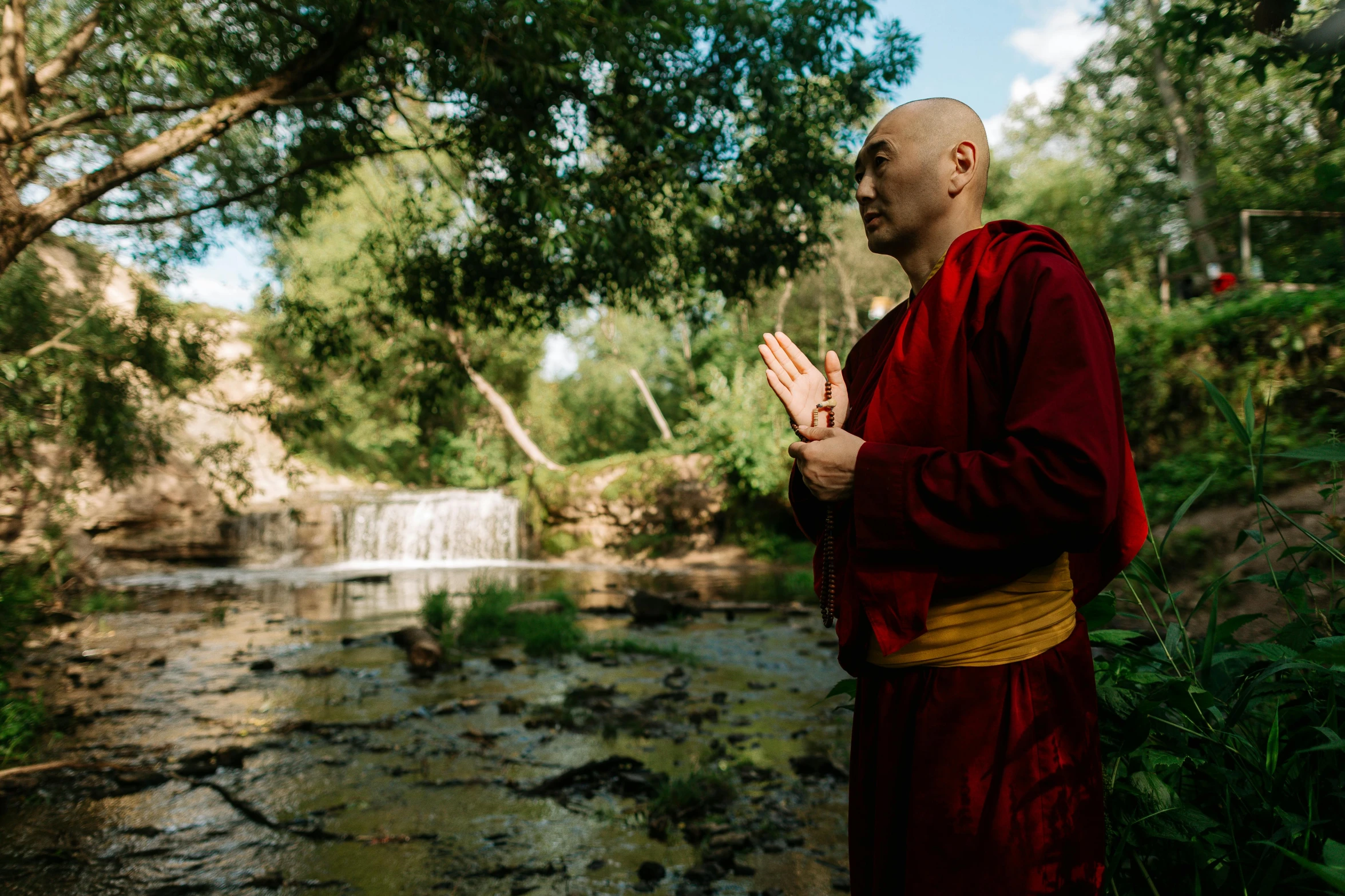 a man in a red robe standing in front of a waterfall, a portrait, inspired by Li Di, unsplash, process art, ukrainian monk, standing gracefully upon a lotus, beautiful sunny day, portrait of bald