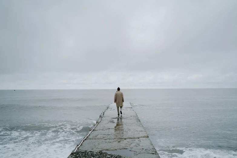 a person standing on a pier next to the ocean, grey sky, ignant, walking towards you, backfacing