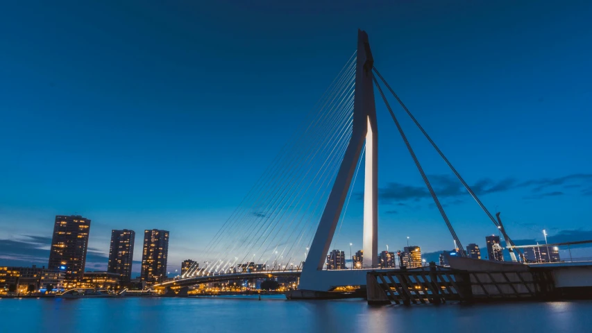 a bridge over a body of water with a city in the background, by Jacob Toorenvliet, pexels contest winner, helmond, blue, in the evening, upright