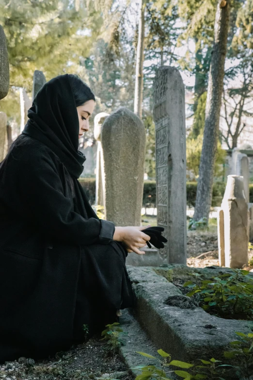 a woman sitting in a cemetery looking at her cell phone, inspired by Modest Urgell, pexels contest winner, wrapped in a black scarf, rip, muslim, from a movie scene