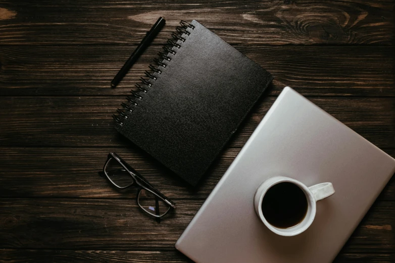 a cup of coffee and a notebook on a wooden table, pexels contest winner, black color scheme, wearing black rimmed glasses, thumbnail, laptops