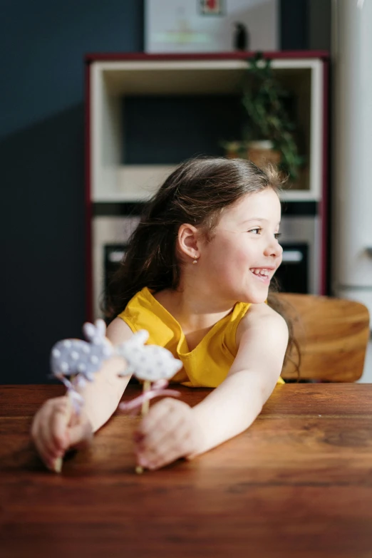a little girl sitting at a table with a flower in her hand, taking control while smiling, professionally assembled, lit from the side, in a kitchen