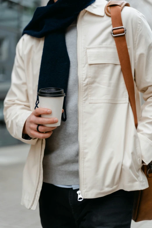 a man walking down a street holding a cup of coffee, by Nina Hamnett, trending on unsplash, cropped shirt with jacket, light cream and white colors, sustainable materials, with two front pockets