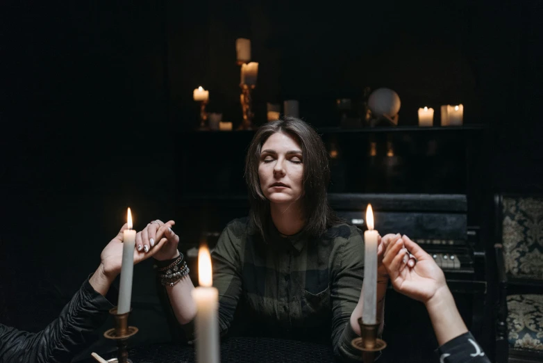 a group of people sitting around a table with candles, a portrait, by Emma Andijewska, female magician, press shot, standing in a dimly lit room, promotional image