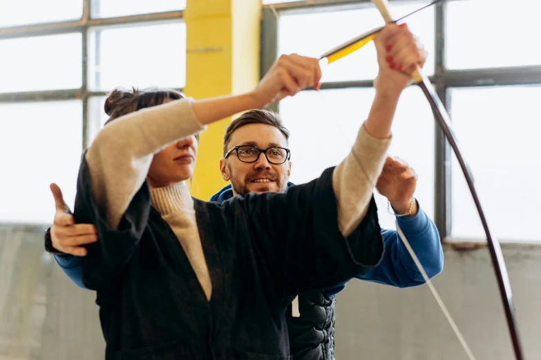 a man and a woman holding a bow and arrow, by Nina Hamnett, pexels contest winner, kinetic art, in a workshop, school class, olafur eliasson, holding a giant flail