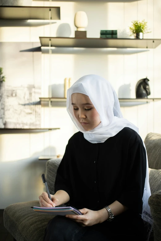 a woman sitting on a couch writing on a piece of paper, inspired by Nazmi Ziya Güran, hurufiyya, wearing white silk hood, architect, malaysian, eva elfie