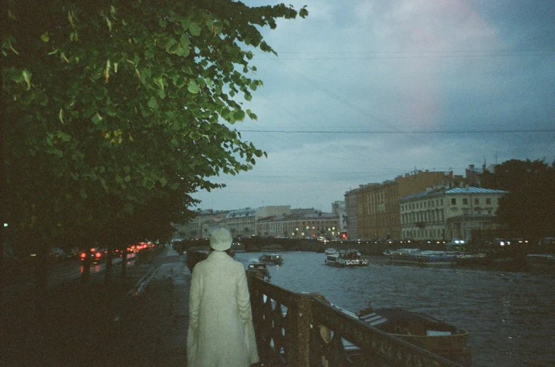 a woman walking down a sidewalk next to a river, an album cover, pexels contest winner, tonalism, saint petersburg, moody : : wes anderson, humid evening, disposable colored camera