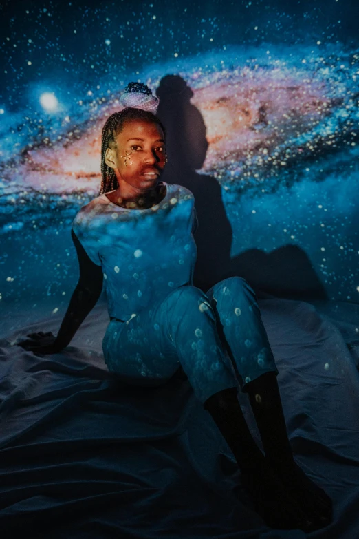a woman sitting on a bed in front of a picture of a galaxy, by Dulah Marie Evans, trending on pexels, afrofuturism, man in dark blue full body suit, water is made of stardust, portrait photo, fullbodysuit