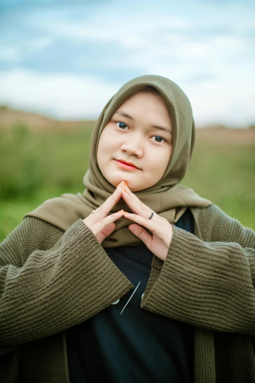 a woman standing in a field with her hands on her chest, hurufiyya, wearing a green sweater, avatar image, casual photography, head scarf