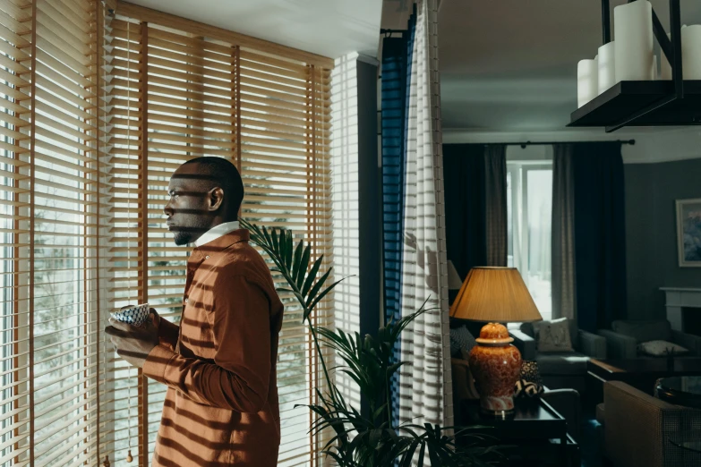 a man standing in front of a window holding a cup, an album cover, pexels contest winner, blind brown man, luxurious environment, soft light through blinds, looking at his phone