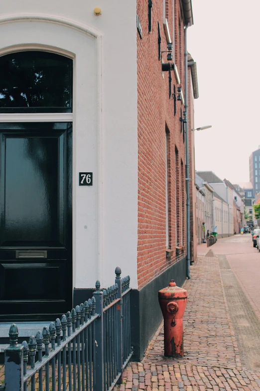 a red fire hydrant sitting in front of a black door, a photo, by Jacob Toorenvliet, unsplash, dutch houses along a river, 70mm film screenshot, street view, brick building