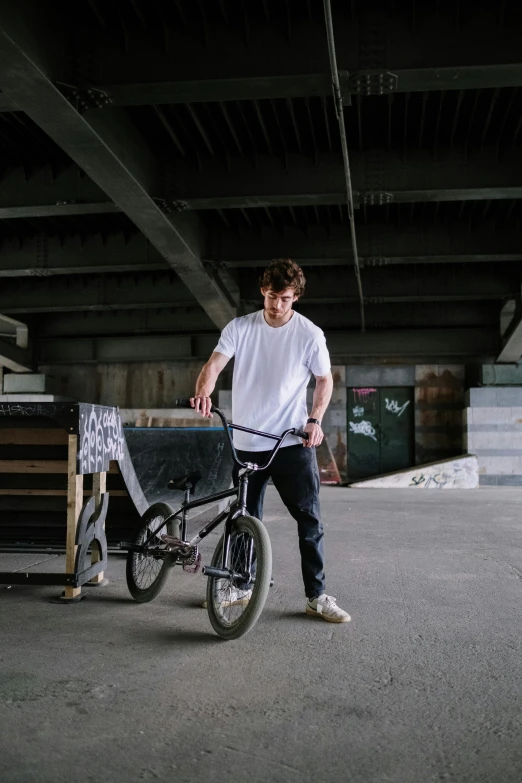 a man standing next to a bicycle in a parking lot, at a skate park, in a warehouse, connor hibbs, promotional image