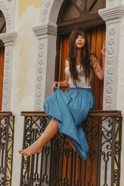 a woman sitting on the balcony of a building, flowing blue skirt, long hair with bangs, promo image, linen