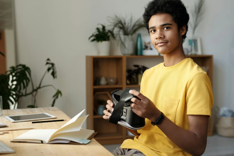 a young man sitting at a desk holding a camera, pexels contest winner, photorealism, black teenage boy, 3d printed, holding a book, gamedev