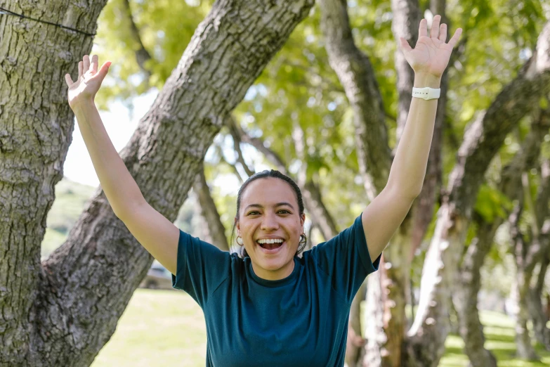 a woman standing next to a tree with her hands in the air, sports photo, she is smiling and excited, an olive skinned, health supporter