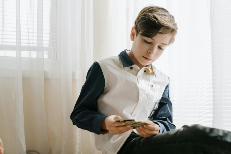 a young boy sitting on a bed looking at a cell phone, pexels contest winner, holding books, wearing a white button up shirt, aged 13, profile pic