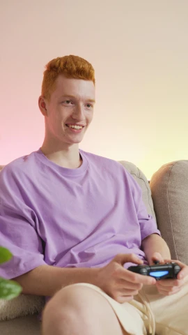 a man sitting on a couch holding a game controller, by Carey Morris, red haired teen boy, purple hue, promo photo, pastel clothing