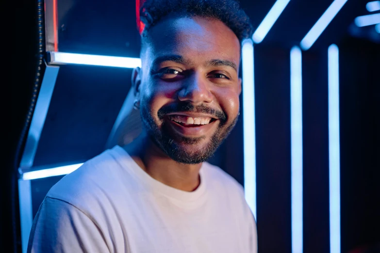 a man in a white shirt smiles at the camera, a character portrait, by Niko Henrichon, pexels contest winner, happening, neon background lighting, mixed race, backroom background, youtube thumbnail
