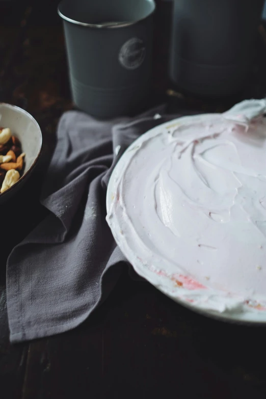 a white plate topped with frosting next to a bowl of nuts, unsplash, process art, white and pink cloth, yogurt, whirlpool, grey