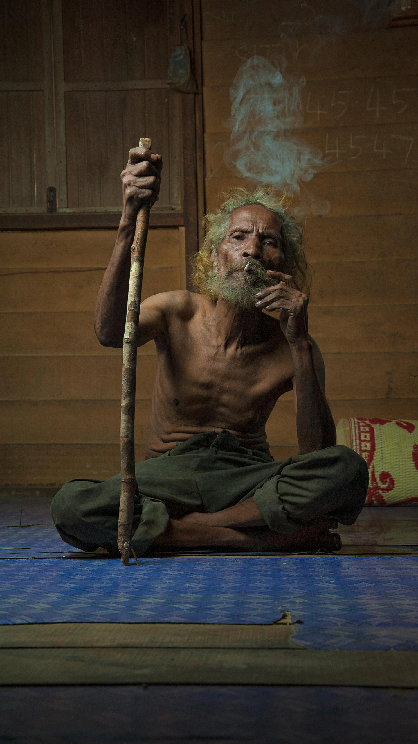 a man sitting on the floor smoking a cigarette, inspired by I Ketut Soki, pexels contest winner, sumatraism, a photo of a disheveled man, holding walking stick, paul barson, padmasana