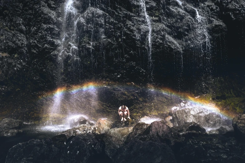 a man standing in front of a waterfall under a rainbow, an album cover, by Beeple, pexels contest winner, magical realism, acid rains. the sacred nipple, grey skies with two rainbows, a beautiful mine, kent monkman