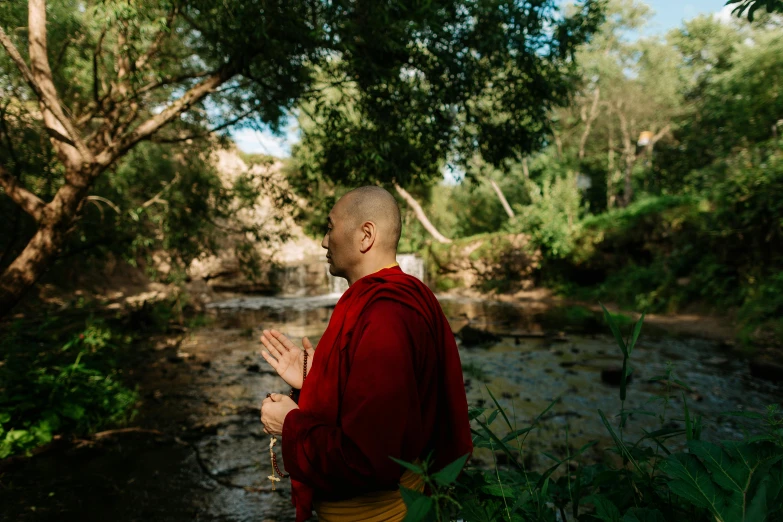 a man in a red robe standing in front of a stream, unsplash, hurufiyya, monk meditate, avatar image, with trees and rivers, behind the scenes photo