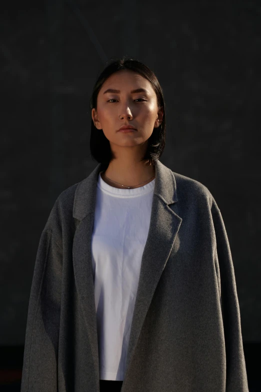 a woman standing in front of a black wall, inspired by Kim Tschang Yeul, unsplash, wearing a grey robe, cropped shirt with jacket, dezeen, south east asian with round face