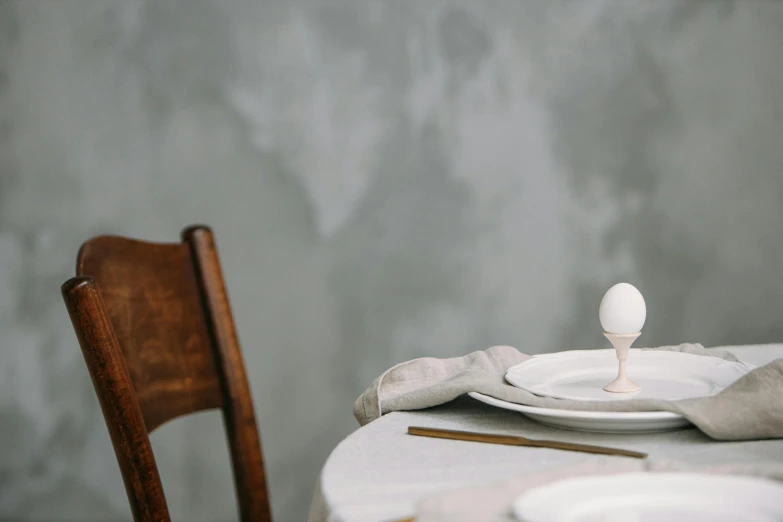 a white plate sitting on top of a table next to a wooden chair, inspired by Alberto Giacometti, unsplash, renaissance, an egg, old kitchen backdrop angled view, linen, looking left