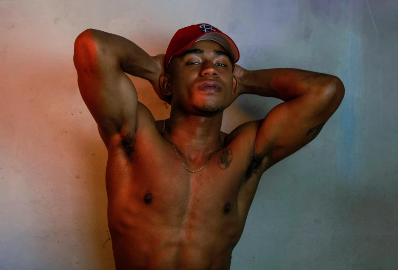 a shirtless man standing in front of a wall, an album cover, by Robbie Trevino, pexels contest winner, wearing a red backwards cap, ( ( dark skin ) ), non binary model, lights off