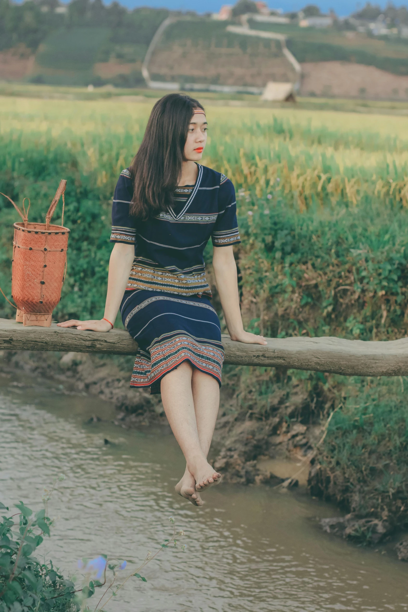 a woman sitting on a log next to a river, inspired by Ruth Jên, pexels contest winner, traditional dress, square, handsome girl, striped