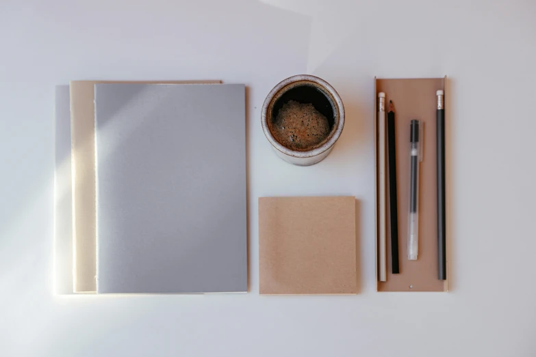 a white table topped with a cup of coffee and a notebook, a minimalist painting, unsplash, gradient brown to silver, pencils, thumbnail, ignant