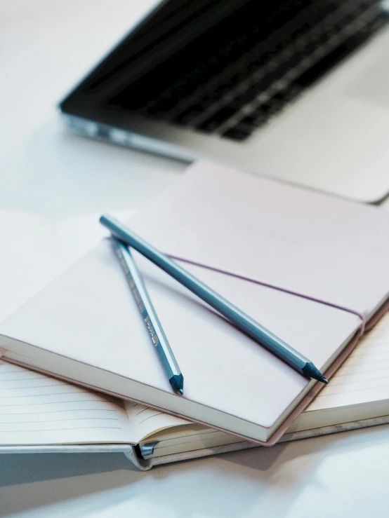 a laptop computer sitting on top of a white desk, pencil and paper, blue ink pen, holding notebook, zoomed in