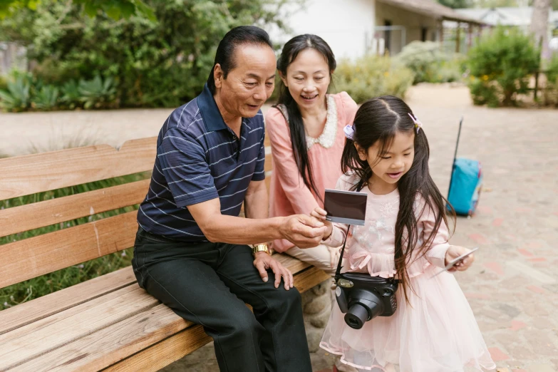 a man sitting on top of a wooden bench next to a little girl, holding a camera, avatar image, asian women, parents watching